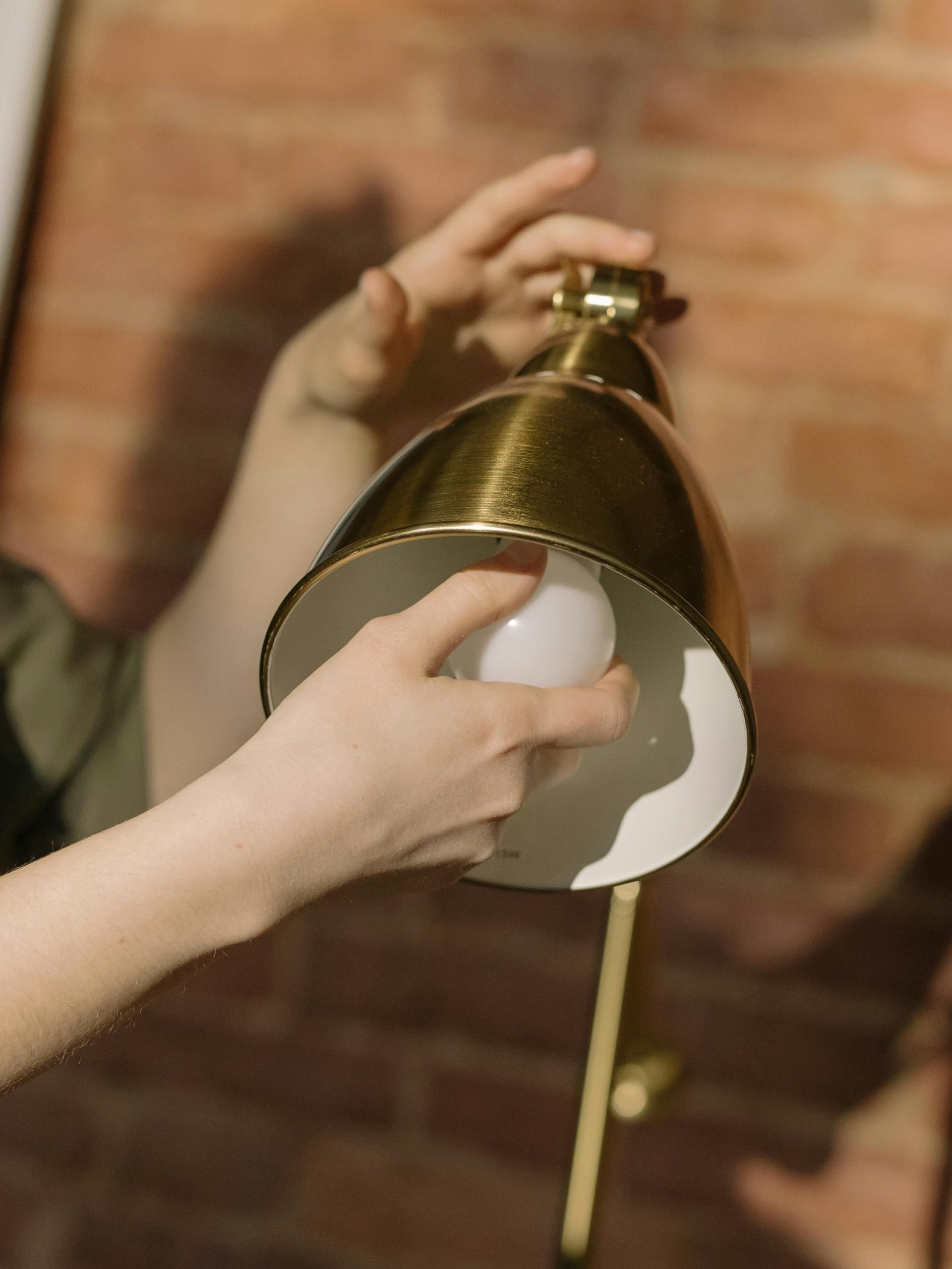 Close-up of hands installing a lightbulb in a brass lamp against a brick wall.